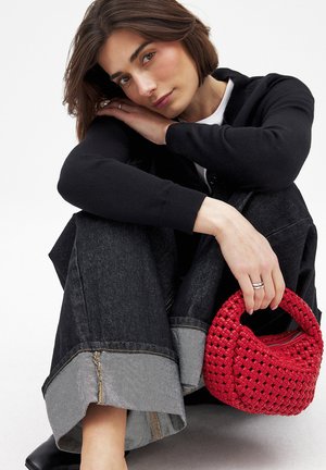 Rote geflochtene Handtasche mit einem runden Griff, zusammen mit dunklen Jeans aus Denim mit umgeschlagenen Säumen und einem schwarzen Cardigan über einem weißen Oberteil.