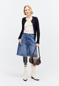 Γυναίκα με κοντά ξανθά μαλλιά που φοράει μαύρη ζακέτα, λευκό τοπ, τζιν σορτς, λευκά μποτάκια, κρατώντας ένα καφέ τσαντάκι, με προσθετικό αριστερό πόδι.