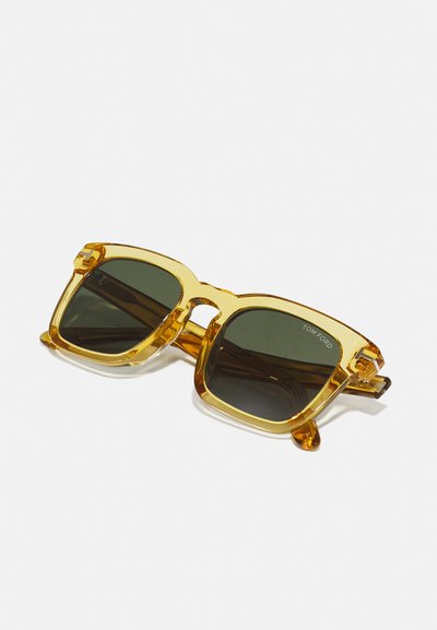 Ruudukujulised prillid, millel on läbipaistev kollane raam ja tumerohelised läätsed. Iseloomulikud jooned on paksud kukkude osad ja sissepressitud logo siseküljel.