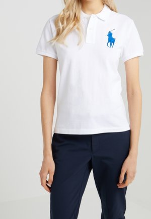 Kobieta ubrana w białą koszulkę polo z krótkim rękawem z niebieskim haftowanym logo gracza polo oraz granatowe spodnie, stojąca na jednolitym tle.