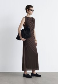 Brauner, drapierter Kleid mit ärmellosem Design, kombiniert mit einer schwarzen Wildleder-Handtasche. Schwarze Slipper runden den Look ab. Flache und glatte Texturen.