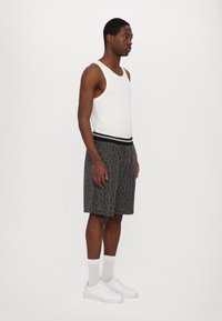 Top de tirantes blanco, pantalones cortos grises con un patrón y una cintura negra, zapatillas blancas y calcetines gris claro. El modelo está de perfil contra un fondo blanco.