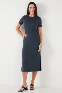 Kvinde står op ad hvid væg iført en tætsiddende, kortærmet, marineblå midi-kjole med sidelomme og sorte hælede sandaler.