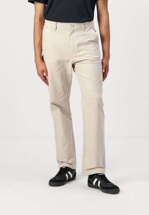 Beige cotton trousers with a straight fit, featuring belt loops, two front pockets, and a side pocket. Worn with black sneakers.