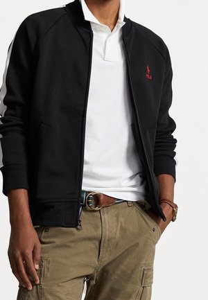 Mann trägt schwarze Zip-up-Jacke über weißem Poloshirt, braunen Ledergürtel und olivgrüne Cargohose, mit einer Hand in der Tasche.