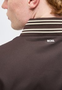 Κοντινό σε λαιμό και αυτί ενός ατόμου που φοράει καφέ μπουφάν με ριγέ γιακά και λογότυπο "BOSS" στην πλάτη.