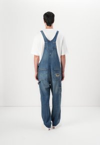 Denim tuinbroeken met verstelbare banden, losse pasvorm en meerdere zakken. Voorzien van geborduurd logo en contrasterende stiksels. Gdragend over een wit shirt.