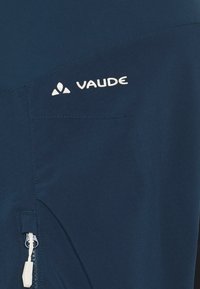 Tumši zila Vaude jaka ar rāvējslēdzēja kabatu, baltu logotipu, gludu tekstūru un kontrastējošiem diegiem jostā.
