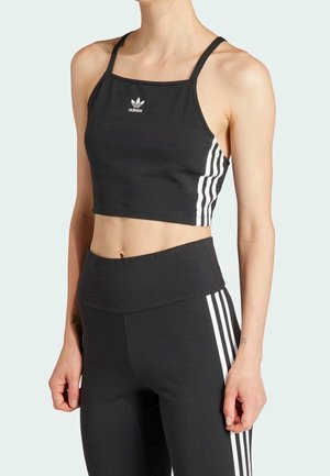 Szczupła osoba ubrana w czarny top Adidas i dopasowane, wysokowysokie legginsy z białymi trzema paskami po bokach na jasnym tle.