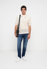 Polo shirt bej din tricot cu mâneci scurte, asortat cu blugi din denim albastri. Modelul poartă o geantă de umăr neagră și teneși albi.