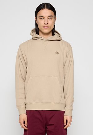 Mann mit langen dunklen Haaren, der einen beigen New Balance Hoodie und burgunderrote Hose trägt, vor einem einfarbigen hellen Hintergrund stehend.