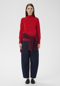 Cămașă roșie cu mânecă lungă, guler înalt și nasturi, asortată cu pantaloni largi din denim albastru. Un fular tricotat burgundy este legat în jurul taliei.