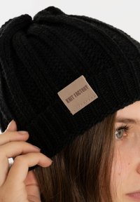Zwarte gebreide beanie met ribbelstructuur, voorzien van een lichtbruine label met de tekst "Knit Factory" gestikt aan de zijkant.