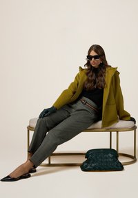 Cappotto verde oliva con cappuccio, maglione nero, pantaloni grigi a quadri, guanti verdi, ballerine nere con cinturino e borsa a tracolla verde intrecciata.