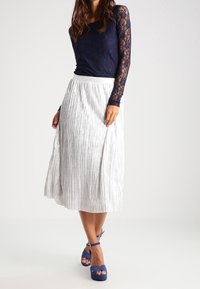 Haut en dentelle bleu marine à manches longues associé à une jupe plissée argentée, présentant une surface texturée, et des sandales bleues à talons hauts avec des lanières.