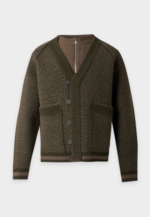 Modèle masculin portant un cardigan vert foncé, un pull zippé marron, un pantalon marron ample et des bottes rouge foncé brillantes avec des lacets rouges, debout sur fond blanc.