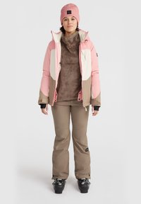 Růžová a béžová izolační bunda přes hnědý fleece top, doplněná béžovými lyžařskými kalhotami a černými lyžařskými botami. Měkká textura, sportovní design.