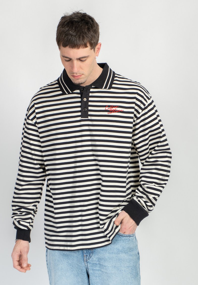 Άνδρας που φοράει μαύρο και άσπρο μπλουζάκι πόλο με οριζόντιες ρίγες και κόκκινο λογότυπο με γράμματα, ανοιχτόχρωμα μπλε τζιν, κοιτάζει προς τα κάτω με το χέρι στην τσέπη.