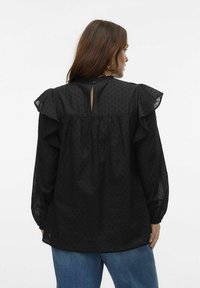 Blusa nera con pois in rilievo, maniche a volant, dettaglio con apertura a chiave sulla schiena e vestibilità ampia, abbinata a jeans in denim blu.