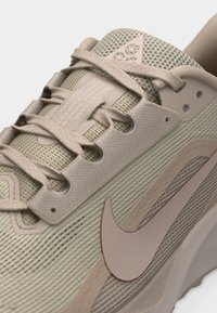 Κοντινή λήψη μπεζ αθλητικού παπουτσιού Nike που δείχνει το υφασμάτινο επάνω μέρος με υφή, τις τρύπες για τα κορδόνια, το λογότυπο Nike και τη μαλακή γλώσσα με τυπωμένο κείμενο και λογότυπο.