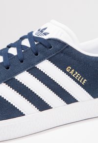 Blue suede sneaker with white leather stripes. Features gold "Gazelle" branding and a textured white rubber sole. Classic lace-up design.