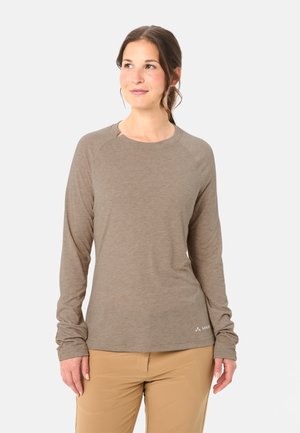 Donna che indossa una maglia beige a maniche lunghe e pantaloni marrone chiaro, che guarda leggermente verso sinistra con uno sfondo grigio neutro.