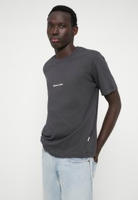 Tumši pelēks kokvilnas T-krekls ar īsām piedurknēm, uz kura krūšu daļas ir vienkāršs balts izšuvēts logo. Mīksta tekstūra un brīvs piegriezums.