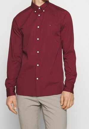 Muž v těsně padnoucí bordó košili na knoflíky s bílými knoflíky a béžových kalhotách stojí před jednoduchým světlým pozadím.