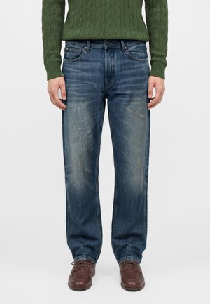 Mand iført falmede blå jeans, brune lædersko og en grøn kabelstrikket sweater, der står mod en ensartet baggrund.
