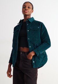 Veste matelassée en velours côtelé teal avec un col, fermeture à boutons sur le devant et deux poches poitrine, portée sur un haut noir et un pantalon sombre.