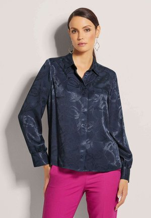 Donkerblauwe blouse met lange mouwen, een subtiel bloemmotief, een kraag met knopen en een gladde, glanzende textuur. Gepaard met roze broeken.