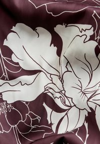 Burgunderfarbener Stoff mit floralem Design in Weißkonturen, der große Blüten und filigrane Blattmuster zeigt. Glatte Textur.