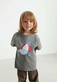 Hallidehall pärlihall T-särk, millel on värviline mütsi graafika punaste, valgete ja siniste aktsentidega, täiendab pruuni riidega püksid.