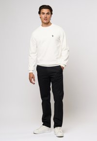 Sweatshirt en coton blanc à manches longues, avec un petit emblème sur la poitrine. Associé à un pantalon noir et des baskets blanches. Coupure mi-longue.