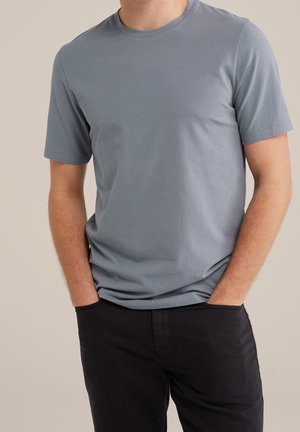 Homme portant un t-shirt gris à manches courtes avec col rond et les mains dans les poches, portant un pantalon noir, devant un fond uni.