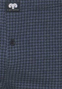 Tissu bleu marine avec un motif pied-de-poule, comportant un seul bouton noir et une petite étiquette logo dans le coin supérieur gauche.