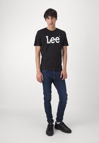 Melna kokvilnas T-kreklā ar baltu "Lee" logotipu, kopā ar tumši ziliem džinsiem un melnām sporta apavām. Vienkāršs, ikdienišķs dizains ar īsām piedurknēm.
