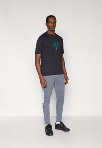 Czarny T-shirt z krótkim rękawem z zielonym napisem "LFC", zestawiony z szarymi spodniami z białymi bocznymi paskami oraz czarnymi butami sportowymi.