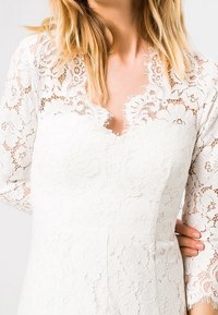 Bílé krajkové šaty s květinovým vzorem, výstřihem do V a dlouhými rukávy. Tkanina má texturovaný vzhled s propracovanými detaily.