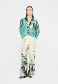 Camicia di seta a fiori in teal, pantaloni a gamba larga bianchi con stampa botanica verde e blu, abbinati a scarpe senza stringhe nere. La modella è in posa eretta.