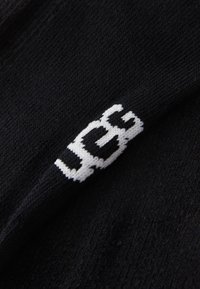 Svart strikket stoff med en hvit brodert UGG-logo i en vinkel, som viser tekstur og fine sømdetaljer.