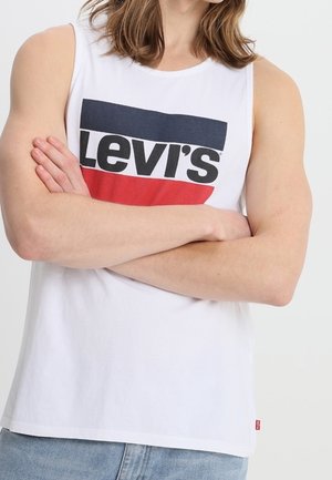 Persona con una camiseta sin mangas blanca de Levi's con el logo en azul marino y rojo, brazos cruzados, combinada con unos vaqueros azul claro.