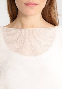 Pull léger de couleur crème avec un design en maille douce et transparente, doté d'un large col et d'une finition texturée.