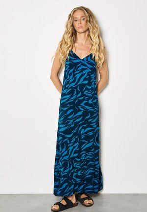 Kobieta o długich falujących blond włosach, ubrana w bezrękawnikową, granatową sukienkę maxi z jasnoniebieskimi abstrakcyjnymi wzorami i czarne sandały, stojąca.