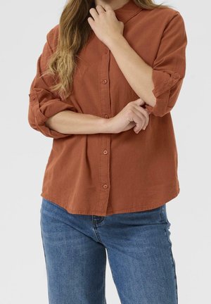 Kvinde med langt bølget hår iført en rustfarvet skjorte med knapper og opkrogede ærmer samt blå jeans, der krydser armene foran kroppen.