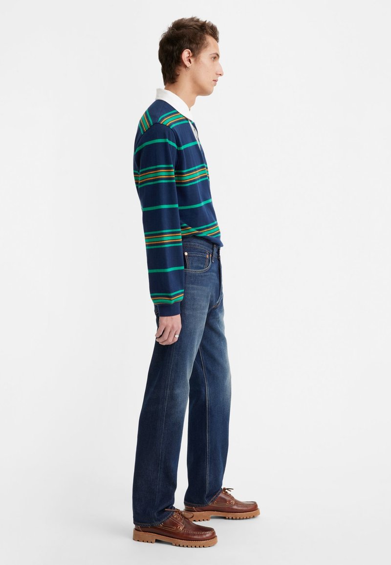 Marineblaues Langarmpolo mit grünen und orangefarbenen Streifen, kombiniert mit dunkelblauen Jeans und braunen Schnürschuhen mit dicken Sohlen.