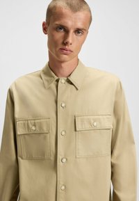 Tan button-up shirt with two front pockets, a collar, and visible stitching details. Features a logo patch on the left pocket.