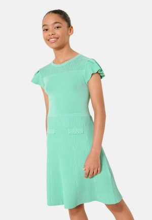 Ung pige smilende, iført en mintgrøn strikket kjole med korte flæseærmer og dekorative forlommer, stående mod en hvid baggrund.