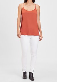 Top senza maniche di colore arancione ruggine con spalline sottili, abbinato a jeans skinny bianchi e stivaletti neri alla caviglia con dettagli in bottone argentato.