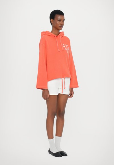 Koralfarbene, übergroße Kapuzenjacke mit Kordelzug, versehen mit einem grafischen Design. Kombiniert mit weißen Shorts und grauen Socken, vor einem schlichten Hintergrund.